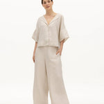 Image of a woman wearing a sand-colored linen top and a matching bottom. The top has a wing collar and the bottom is long and flowy. The purpose of the image is to highlight the clothing by NAOS because it's available for sale at EVAMAIA boutique in Studio City, CA.