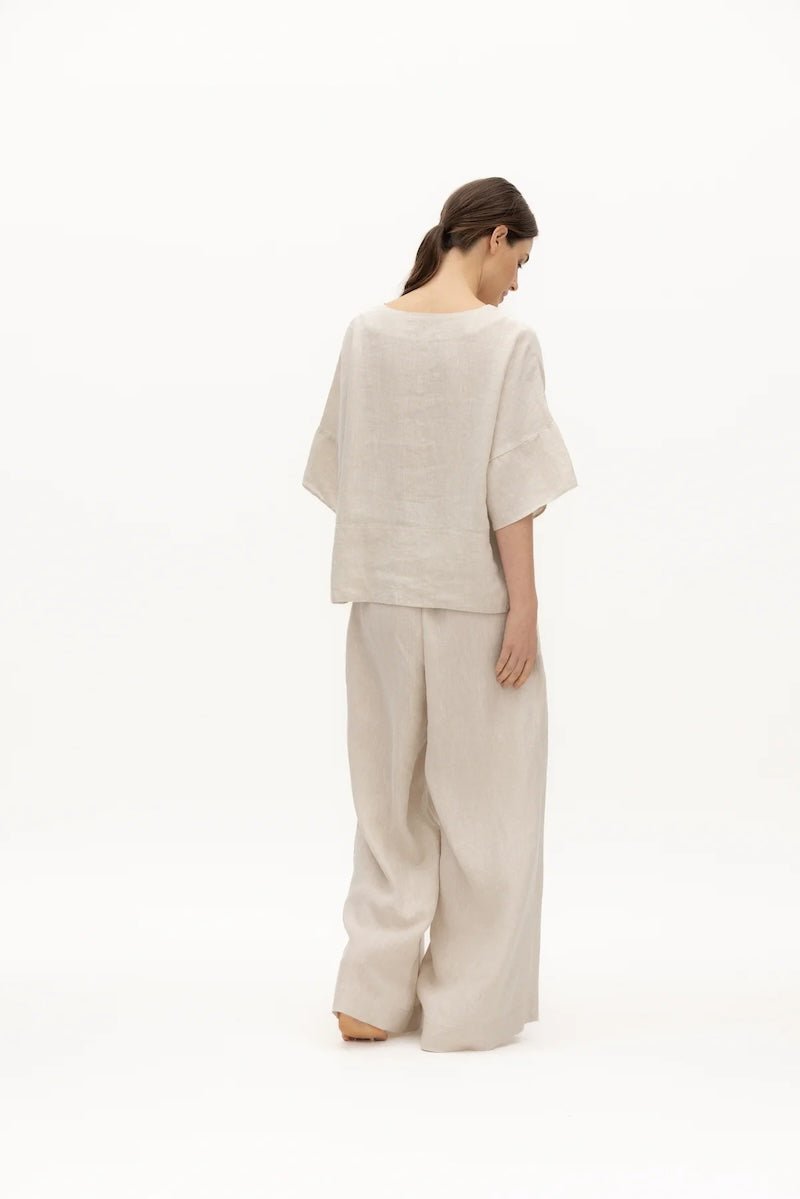 Image of a woman wearing a sand-colored linen top and a matching bottom. The top has a wing collar and the bottom is long and flowy. The purpose of the image is to highlight the clothing by NAOS because it's available for sale at EVAMAIA boutique in Studio City, CA.