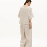 Image of a woman wearing a sand-colored linen top and a matching bottom. The top has a wing collar and the bottom is long and flowy. The purpose of the image is to highlight the clothing by NAOS because it's available for sale at EVAMAIA boutique in Studio City, CA.