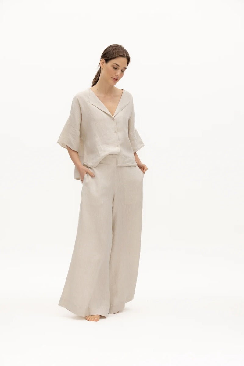 Image of a woman wearing a sand-colored linen top and a matching bottom. The top has a wing collar and the bottom is long and flowy. The purpose of the image is to highlight the clothing by NAOS because it's available for sale at EVAMAIA boutique in Studio City, CA.