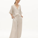 Image of a woman wearing a sand-colored linen top and a matching bottom. The top has a wing collar and the bottom is long and flowy. The purpose of the image is to highlight the clothing by NAOS because it's available for sale at EVAMAIA boutique in Studio City, CA.