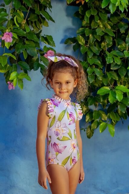 A one-piece swimsuit for girls with a soft lilac base and vibrant orchid print, featuring ruffled cap sleeves and a high neckline.