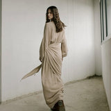 Woman in a long, flowing beige dress standing in a minimalistic room with white walls and concrete floor.