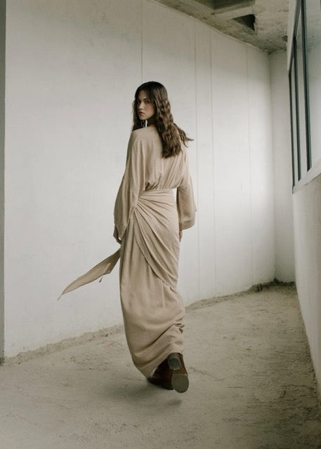 Woman in a long, flowing beige dress standing in a minimalistic room with white walls and concrete floor.
