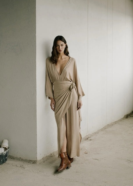 Woman wearing a beige dress leaning against a white wall.