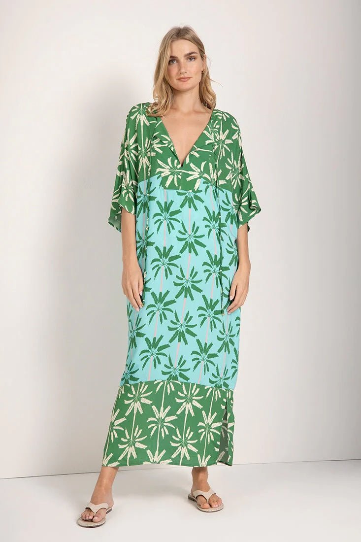 Woman wearing a long cover-up dress with a green and blue palm tree print. She's looking towards the camera and standing in front of a white background. The purpose of this image is to highlight the dress.