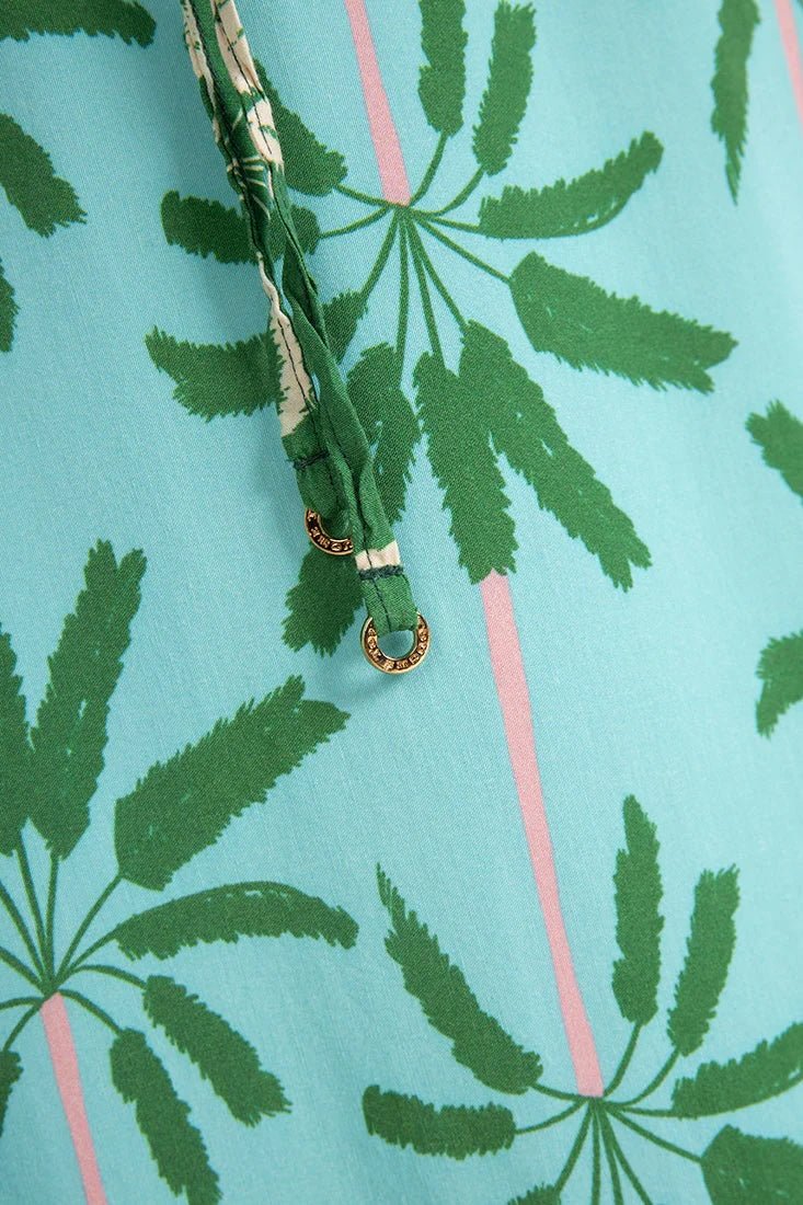 Close up image of a woman wearing a long cover-up dress with a green and blue palm tree print. This close-up just shows the straps, which have a gold detail on the end. The purpose of this image is to highlight the dress.