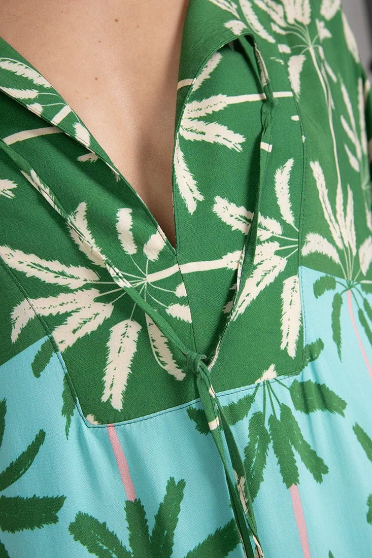 Close up image of a woman wearing a long cover-up dress with a green and blue palm tree print. This close-up just shows the neckline. The purpose of this image is to highlight the dress.