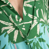 Close up image of a woman wearing a long cover-up dress with a green and blue palm tree print. This close-up just shows the neckline. The purpose of this image is to highlight the dress.
