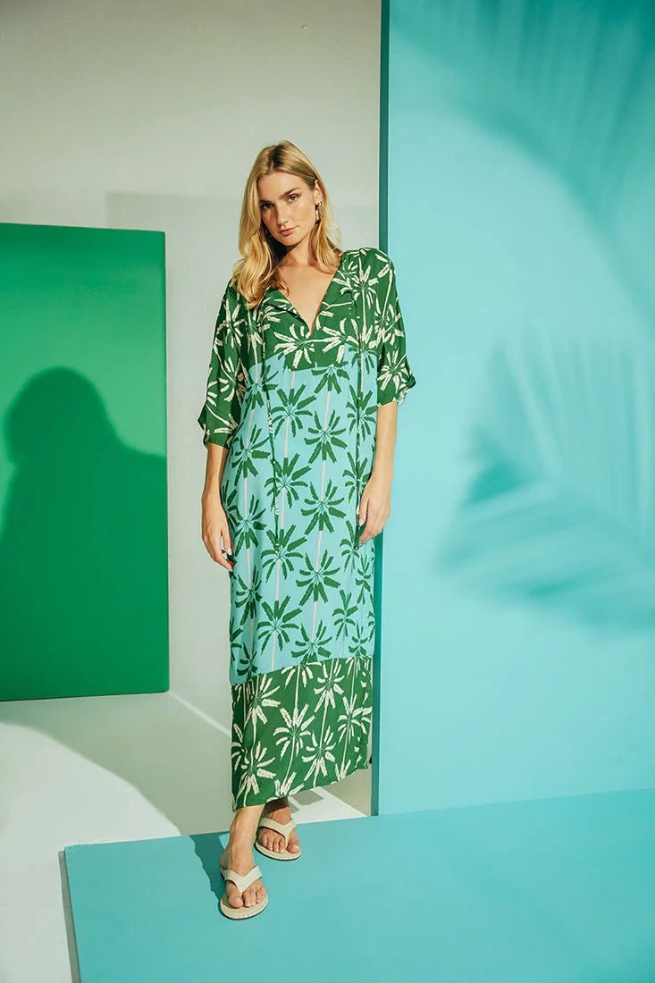 Woman wearing a long cover-up dress with a green and blue palm tree print. She's looking towards the camera and standing in front of a white, teal and blue background. The purpose of this image is to highlight the dress.
