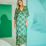 Woman wearing a long cover-up dress with a green and blue palm tree print. She's looking towards the camera and standing in front of a white, teal and blue background. The purpose of this image is to highlight the dress.