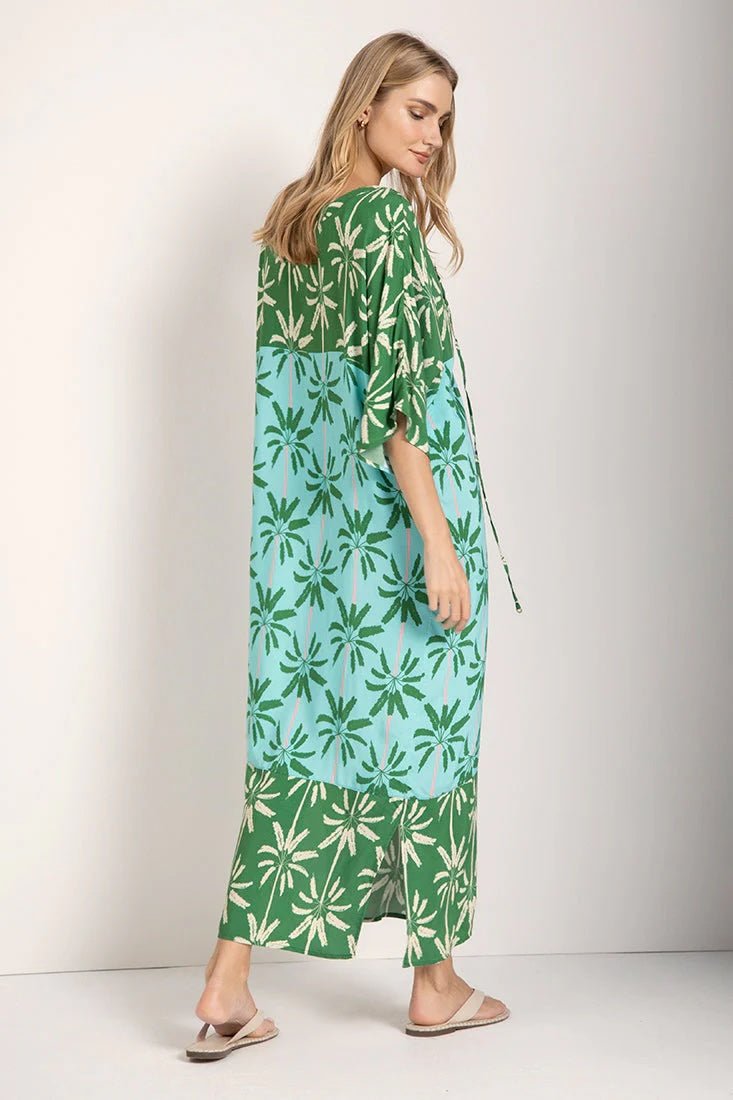 Woman wearing a long cover-up dress with a green and blue palm tree print. She's looking down and to the side. Her body is turned away from the camera. She's standing against a white background. The purpose of this image is to highlight the dress.