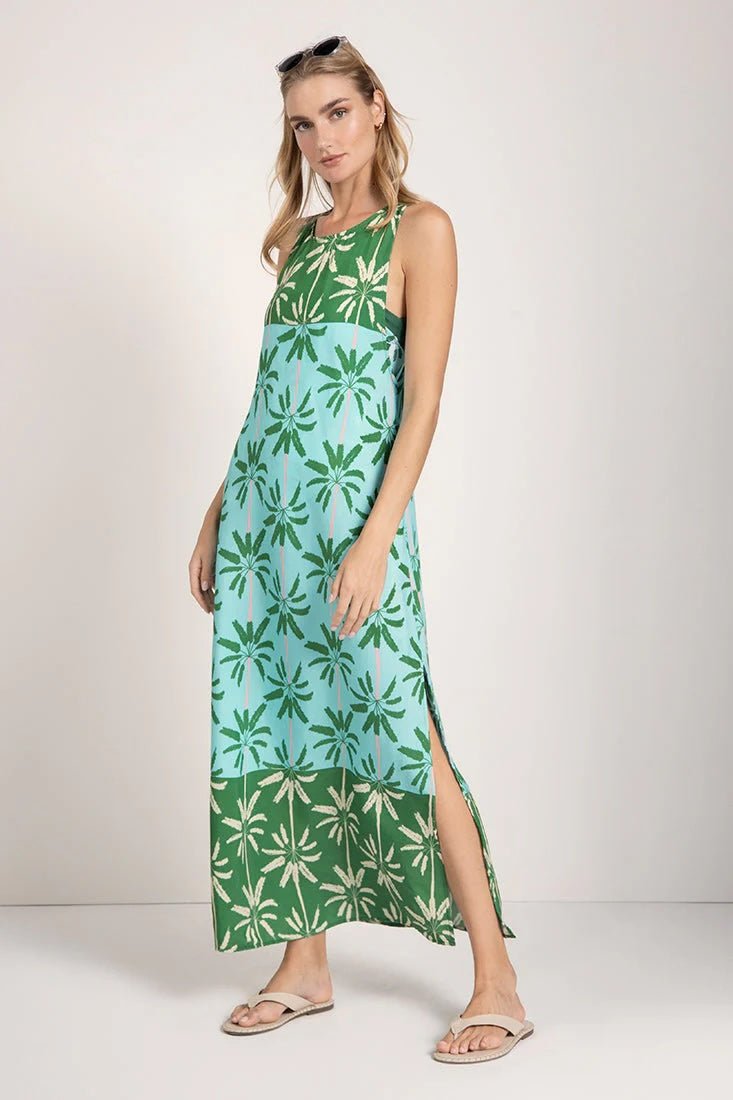 Woman wearing a sleeveless long cover-up dress with a green and blue palm tree print. She's looking towards the camera and standing in front of a white background. The purpose of this image is to highlight the dress.