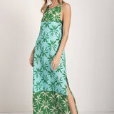 Woman wearing a sleeveless long cover-up dress with a green and blue palm tree print. She's looking towards the camera and standing in front of a white background. The purpose of this image is to highlight the dress.