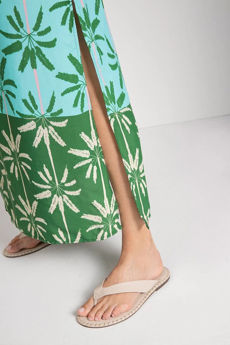 Close up image of a woman wearing a cover-up dress with a green and blue palm tree print. She's standing on a white background. The purpose of this image is to highlight slit on the bottom of the dress.