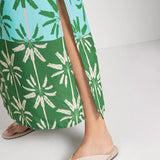 Close up image of a woman wearing a cover-up dress with a green and blue palm tree print. She's standing on a white background. The purpose of this image is to highlight slit on the bottom of the dress.