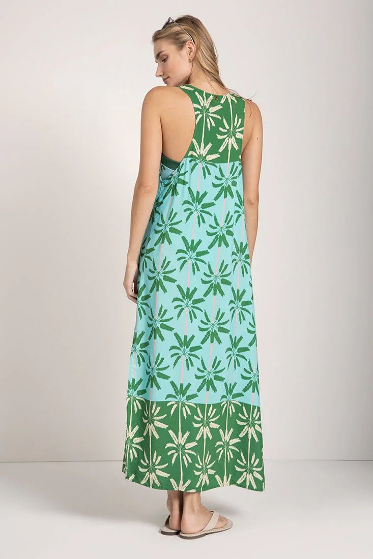 Woman wearing a sleeveless long cover-up dress with a green and blue palm tree print. She's looking to the side and standing against a white background. The purpose of this image is to highlight the dress.