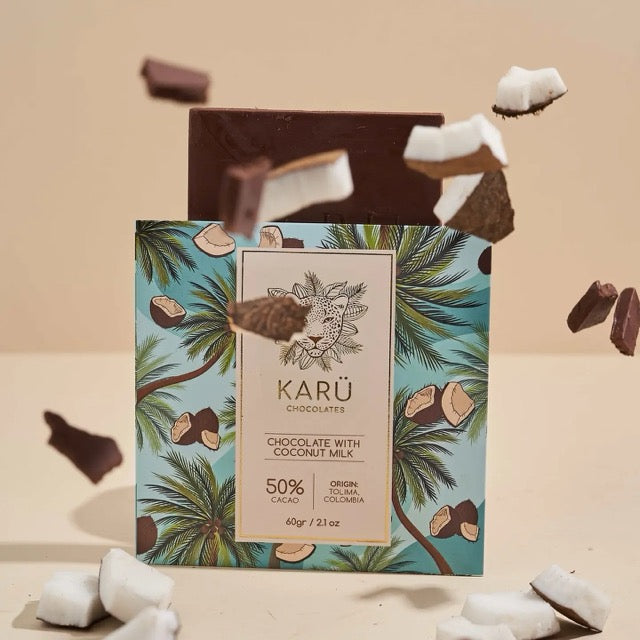 A chocolate bar with coconut milk and '50% CACAO' text on it, with palm trees on the packaging. Coconut pieces are scattered around, and the bar has a sticker with the brand 'Karü'.