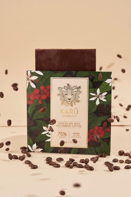 A chocolate bar with '75% Colombian Coffee' text on it, presented on a card with a floral pattern. Chocolate beans are scattered around, and the bar has a sticker with the brand 'Karü'.