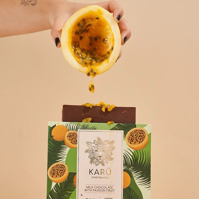A person holding a half of passionfruit above a bar of Keru Chocolates passionfruit flavored chocolate with a background of a box packaged with tropical fruit graphics.
