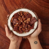 Hands holding half a coconut that is being used as a bowl with pieces of granola and pieces of chocolate.
