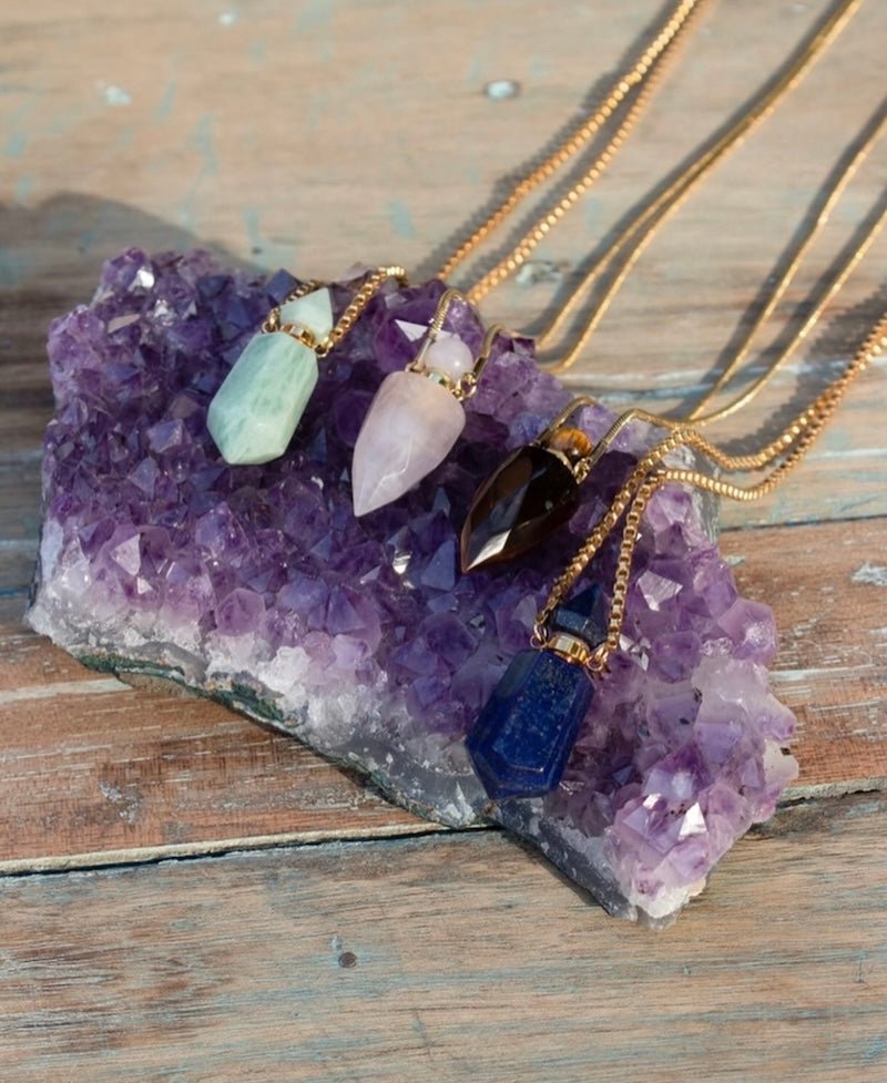 Image of perfume bottles made out of stone sitting on a raw amethyst stone. The purpose of this image is to highlight the perfume bottles, which can be worn as necklaces. They are made by Pajarolimon and sold at EVAMAIA boutique.