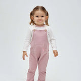 A toddler wearing a pink seahorse overall with white long-sleeved top underneath. The overall has front pockets and embroidered seahorse detailing.
