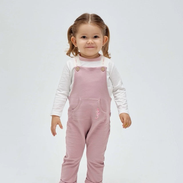 A toddler wearing a pink seahorse overall with white long-sleeved top underneath. The overall has front pockets and embroidered seahorse detailing.