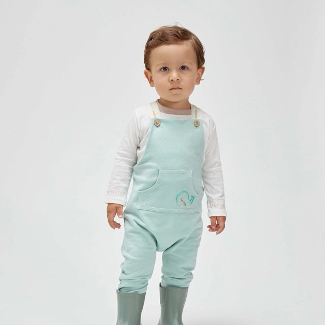 A toddler wearing a light blue cotton overall with a embroidered whale on the pocket, paired with a white long-sleeved shirt and green boots.