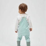 A toddler wearing a light blue cotton overall with a embroidered whale on the pocket, paired with a white long-sleeved shirt and green boots.