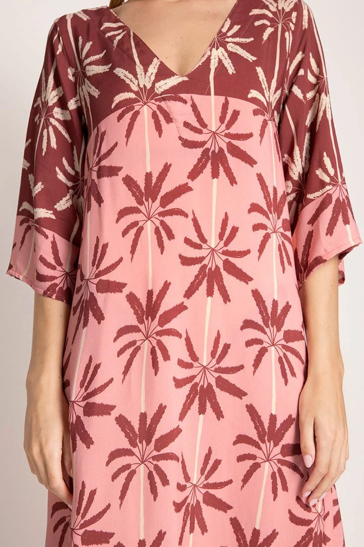 Close up image of a woman wearing a long cover-up dress with a pink and tan palm tree print. We just see her torso. She's standing in front of a white background. The purpose of this image is to highlight the dress.