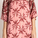 Close up image of a woman wearing a long cover-up dress with a pink and tan palm tree print. We just see her torso. She's standing in front of a white background. The purpose of this image is to highlight the dress.