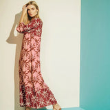 Woman wearing a long cover-up dress with a pink and tan palm tree print. She's looking towards the camera and standing in front of a turquoise and white background. The purpose of this image is to highlight the dress.