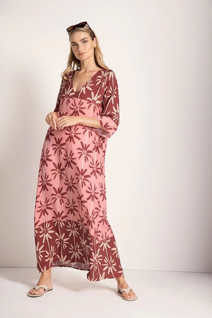 Woman wearing a long cover-up dress with a pink and tan palm tree print. She's looking towards the camera and standing in front of a white background. The purpose of this image is to highlight the dress.