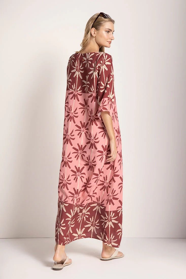 Woman wearing a long cover-up dress with a pink and tan palm tree print. She's looking towards the side and giving her back to the camera. She's standing in front of a white background. The purpose of this image is to highlight the dress.
