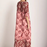 Woman wearing a long cover-up dress with a pink and tan palm tree print. She's looking towards the side and giving her back to the camera. She's standing in front of a white background. The purpose of this image is to highlight the dress.