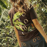 Person wearing a brown t-shirt with 'Colombia' written as well as banana plants and coffee plant design on the back, standing in a forest.