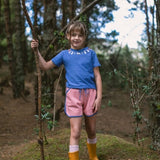 Child in a blue shirt and pink shorts standing in a forest with yellow rain boots.