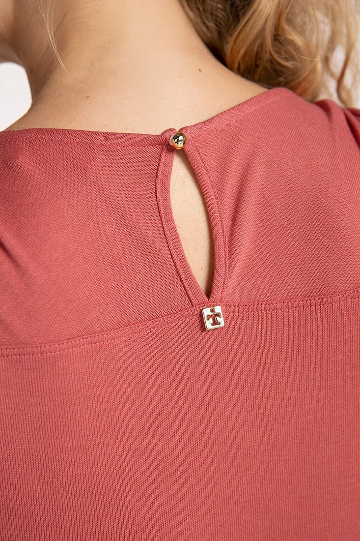 Close up image of a woman wearing a rose colored pajama set. The top has a closure detail on the back. She's standing in front of a white wall. The purpose of this image is to highlight the gold closure detail.