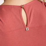 Close up image of a woman wearing a rose colored pajama set. The top has a closure detail on the back. She's standing in front of a white wall. The purpose of this image is to highlight the gold closure detail.