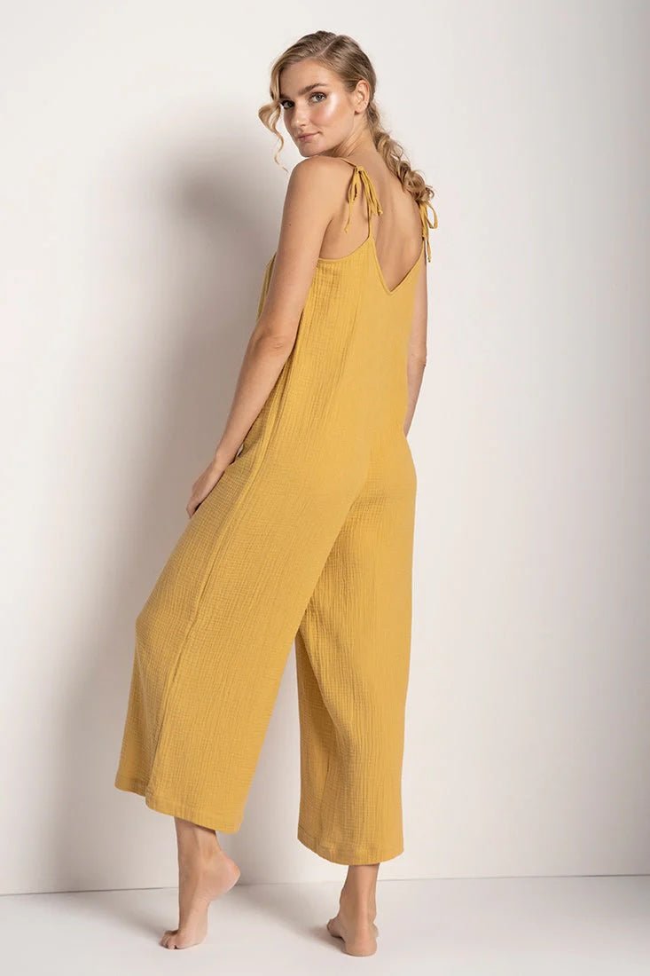 Woman wearing a rusted yellow jumpsuit with tie detail on the sleeves. She's standing in front of a white background and looking to the camera while showing the back of the jumpsuit. The purpose of this image is to highlight the jumpsuit.
