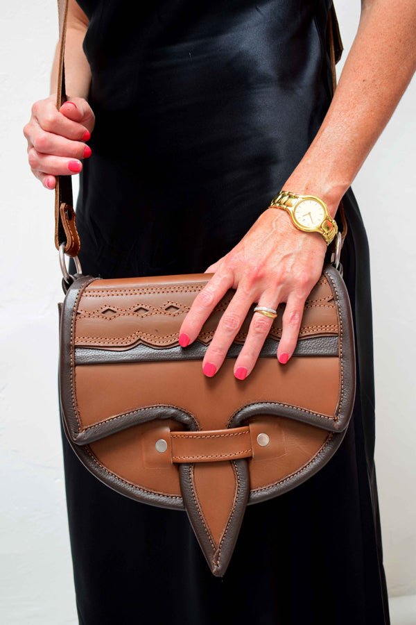 A two-toned brown leather crossbody saddle bag with multiple pockets and hardware accents, handmade in Colombia.