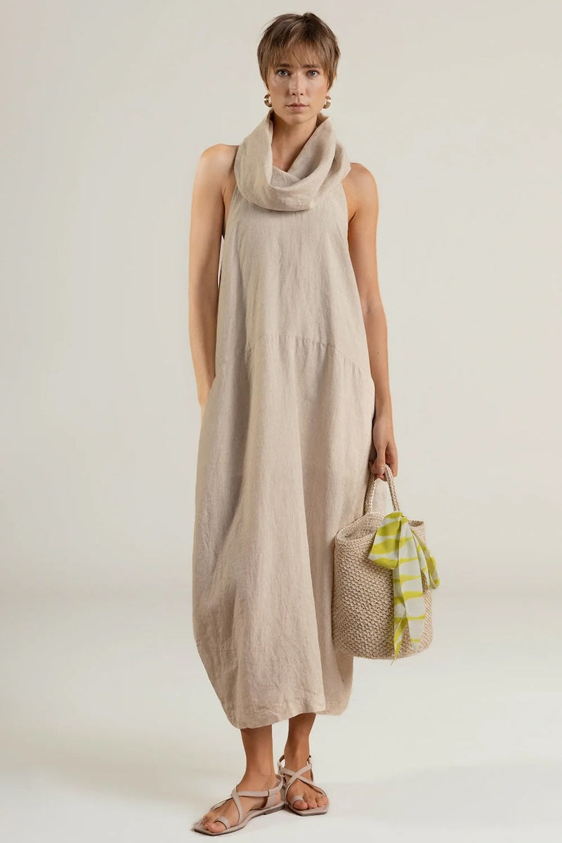 Woman wearing a beige sleeveless dress holding a woven bag on a white background.