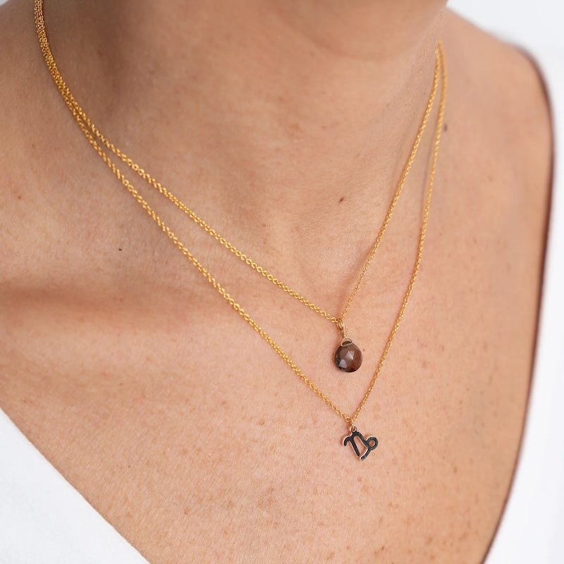 Close up image of a woman's neckline. She's wearing two necklaces. One has a charm of a Capricorn zodiac sign and the other has a smoky quartz stone. The purpose of this image is to highlight the necklaces, which are made in Colombia and sold in Studio City, at EVAMAIA boutique.