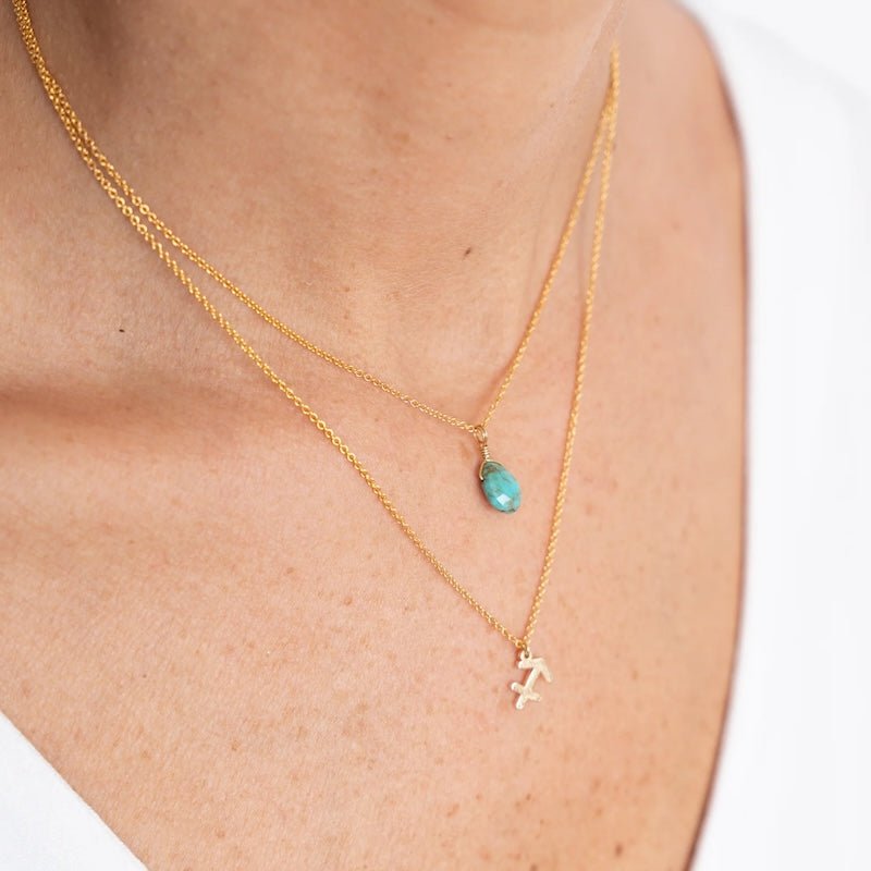 Close up image of a woman's neckline. She's wearing two necklaces. One has a charm of a Scorpio zodiac sign and the other has a Turquoise stone. The purpose of this image is to highlight the necklaces, which are made in Colombia and sold in Studio City, at EVAMAIA boutique.