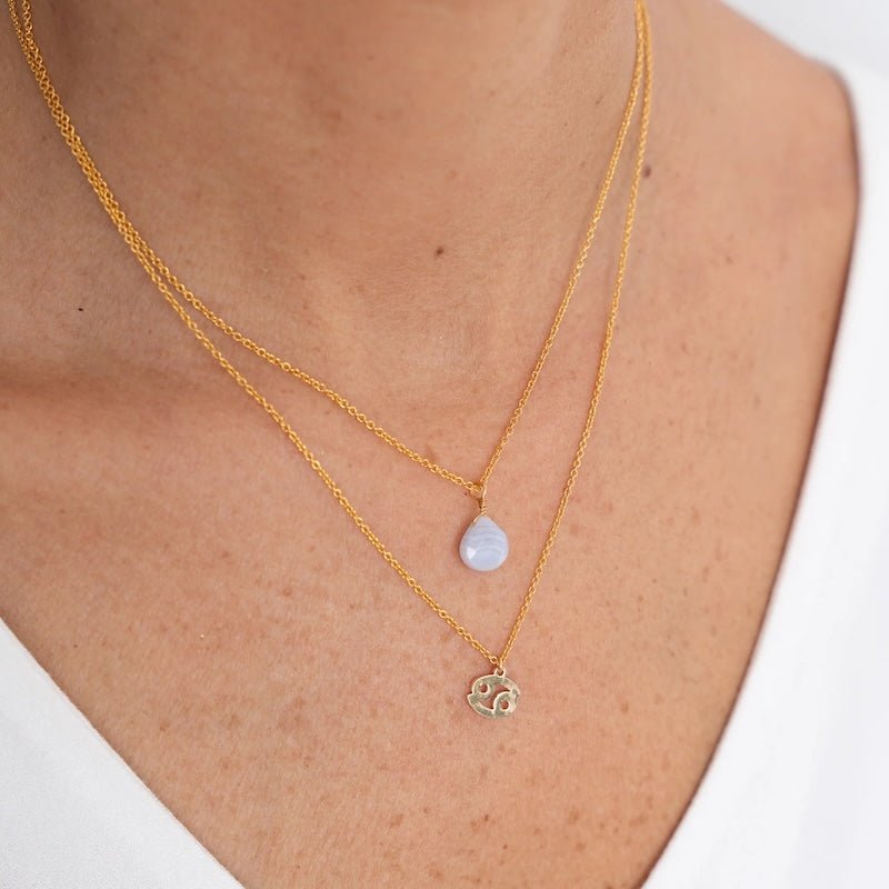 Close up image of a woman's neckline. She's wearing two necklaces. One has a charm of a Cancer zodiac sign and the other has a Blue Ribbon Agate stone. The purpose of this image is to highlight the necklaces, which are made in Colombia and sold in Studio City, at EVAMAIA boutique.