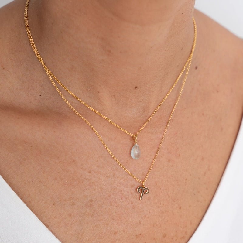 Close up image of a woman's neckline. She's wearing two necklaces. One has a charm of an Aries zodiac sign and the other has an aquamarine stone. The purpose of this image is to highlight the necklaces, which are made in Colombia and sold in Studio City, at EVAMAIA boutique.