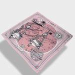 Zodiac Scarf for Libra made by Plissé. Image of scales. It is on top of a astrological background. Details like a white ecliptic, sun, and moon. The scarf is pink with white details floating on plain background.