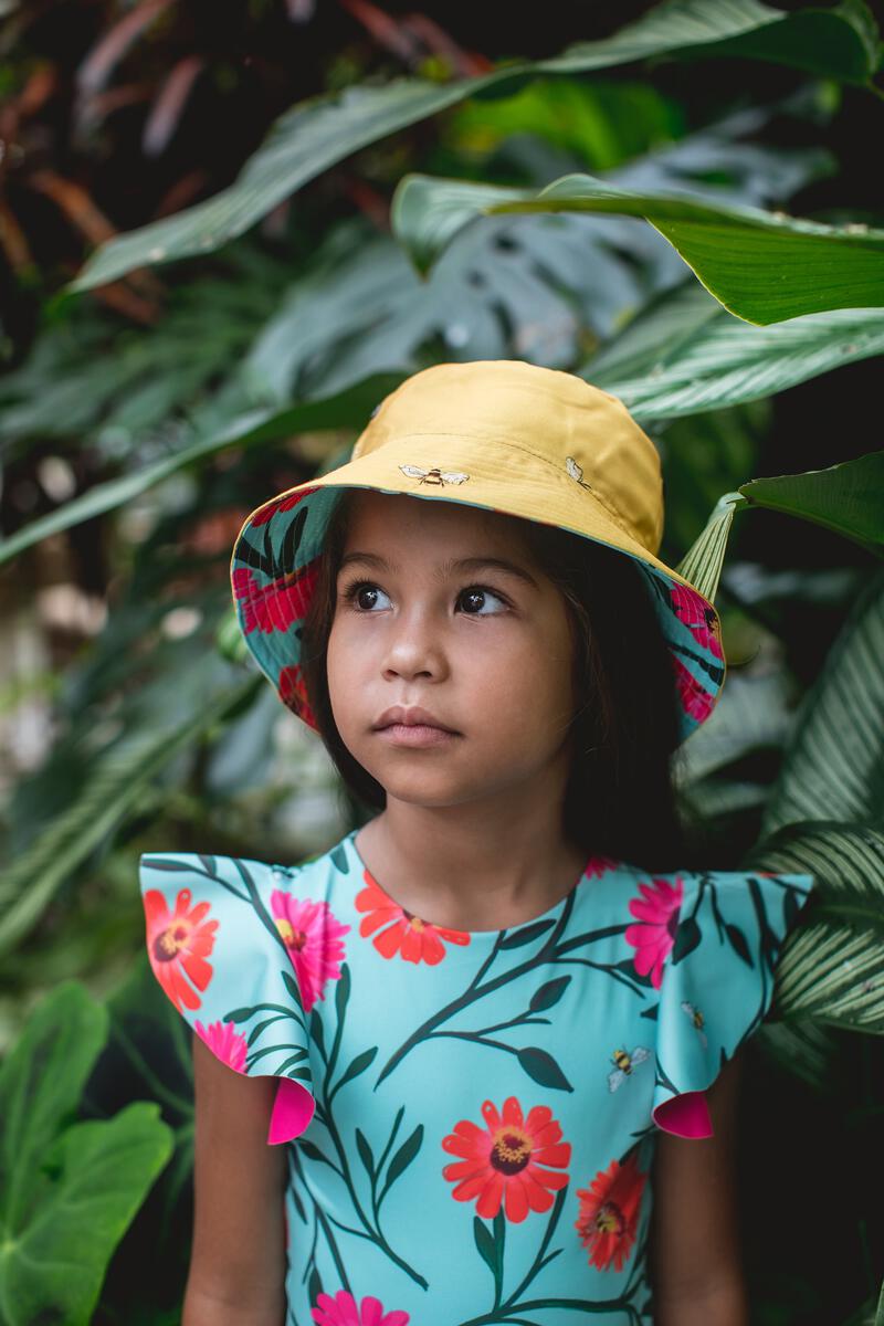Aqua colored Girls' Dayana One Piece with a flower and bee pattern. She is wearing a matching reversible bucket hat in a forest like background.