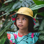 Aqua colored Girls' Dayana One Piece with a flower and bee pattern. She is wearing a matching reversible bucket hat in a forest like background.